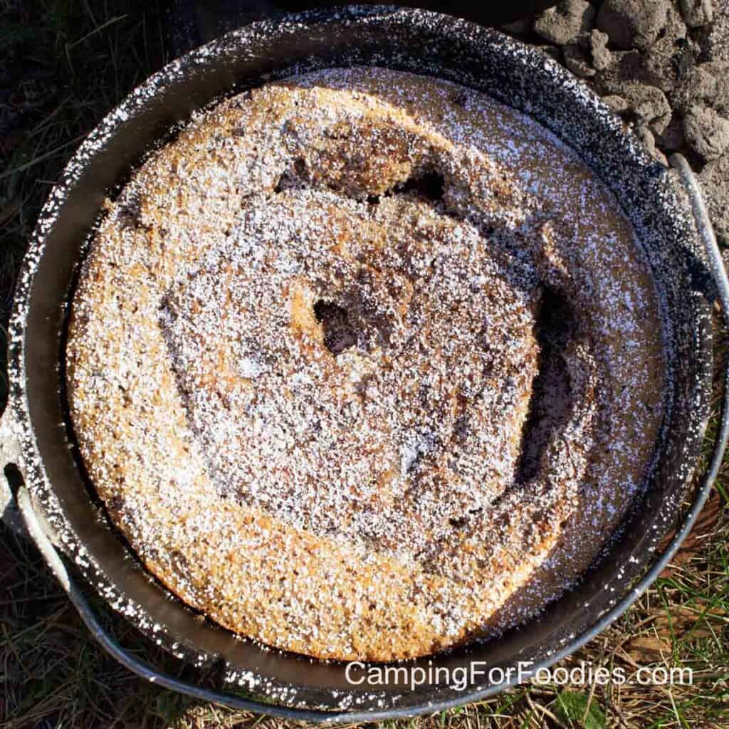 Dutch Oven Coffee Cake by CampingForFoodies features a black cast iron camp Dutch oven filled with a golden brown coffee cake sprinkled with powdered sugar ready to be sliced and served at the campsite.