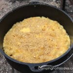 Dutch Oven Mac And Cheese Camping Recipe by CampingForFoodies features a black cast iron camp Dutch oven pot sitting on the ground and filled with creamy macaroni and cheese topped with a golden brown crumb topping ready to be served at the campsite.