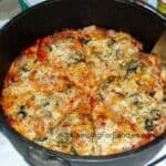 Dutch Oven Pizza Camping Recipe by CampingForFoodies features a black cast iron camp Dutch oven filled with a sliced hot pizza with golden brown cheese, black olives, green peppers and a red tomato sauce ready to be served at the campsite.