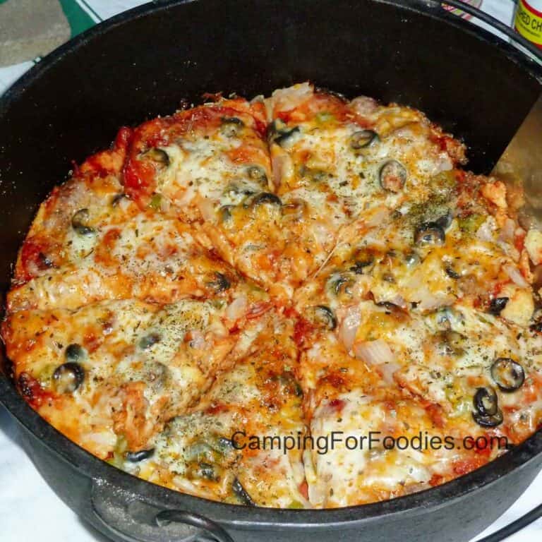 Dutch Oven Pizza Camping Recipe by CampingForFoodies features a black cast iron camp Dutch oven filled with a sliced hot pizza with golden brown cheese, black olives, green peppers and a red tomato sauce ready to be served at the campsite.