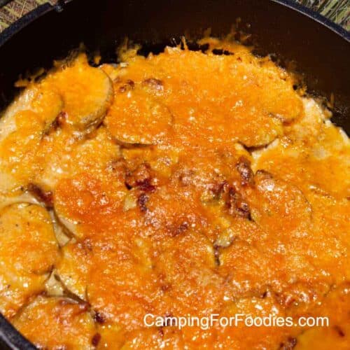 Dutch Oven Potatoes Au Gratin by CampingForFoodies features a black cast iron camp Dutch oven filled with yellow, golden brown melted cheese and a creamy sauce with potato slices.