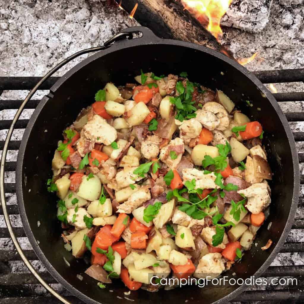 Easy Dutch Oven Chicken And Potatoes by CampingForFoodies features a black cast iron camp Dutch oven sitting on a grate in a campfire ring with a wooden log burning in the background, the pot is filled with a camping dinner of chicken, potatoes and carrots all topped with bright green parsley.