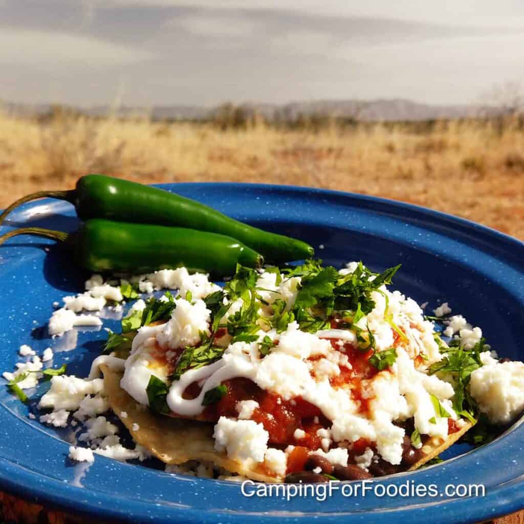 New Mexican Huevos Rancheros by CampingForFoodies features a blue camping plate filled with a golden brown fried tortilla topped with beans, a fried egg, red salsa, drizzle of sour cream, crumbled queso fresco cheese and bright green cilantro with a background of mountains under a cloudy sky.