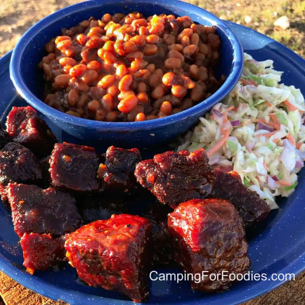 Poor Man Burnt Ends by CampingForFoodies features a blue camping plate filled with wet burnt ends slathered in BBQ sauce, next to fresh coleslaw and a bowl filled with a side of baked beans.
