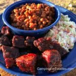 Poor Man Burnt Ends by CampingForFoodies features a blue camping plate filled with wet burnt ends slathered in BBQ sauce, next to fresh coleslaw and a bowl filled with a side of baked beans.