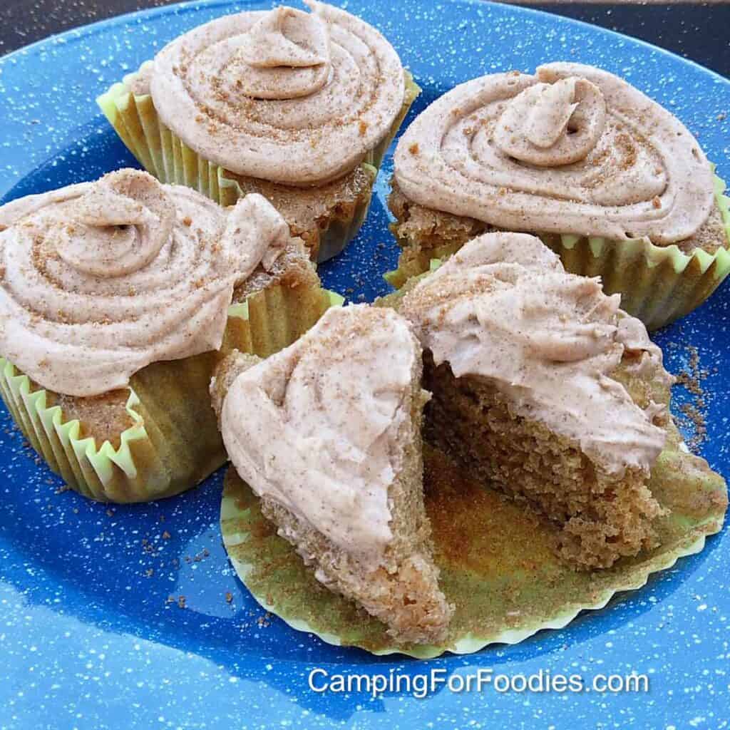 Cinnamon Cupcake Recipe by CampingForFoodies features four cinnamon cupcakes on a blue camping plate. The light brown frosting is swirled on top of each cupcake and topped with brown sugar and cinnamon sprinkles. One cupcake is cut in half showing the moist cupcake center.