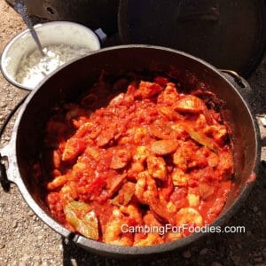 Dutch Oven Jambalaya Recipe by CampingForFoodies features a cast iron camping Dutch oven filled with chicken, sausage and shrimp jambalaya alongside a pot of white rice in the background and a cast iron lid resting against a camping fire ring.