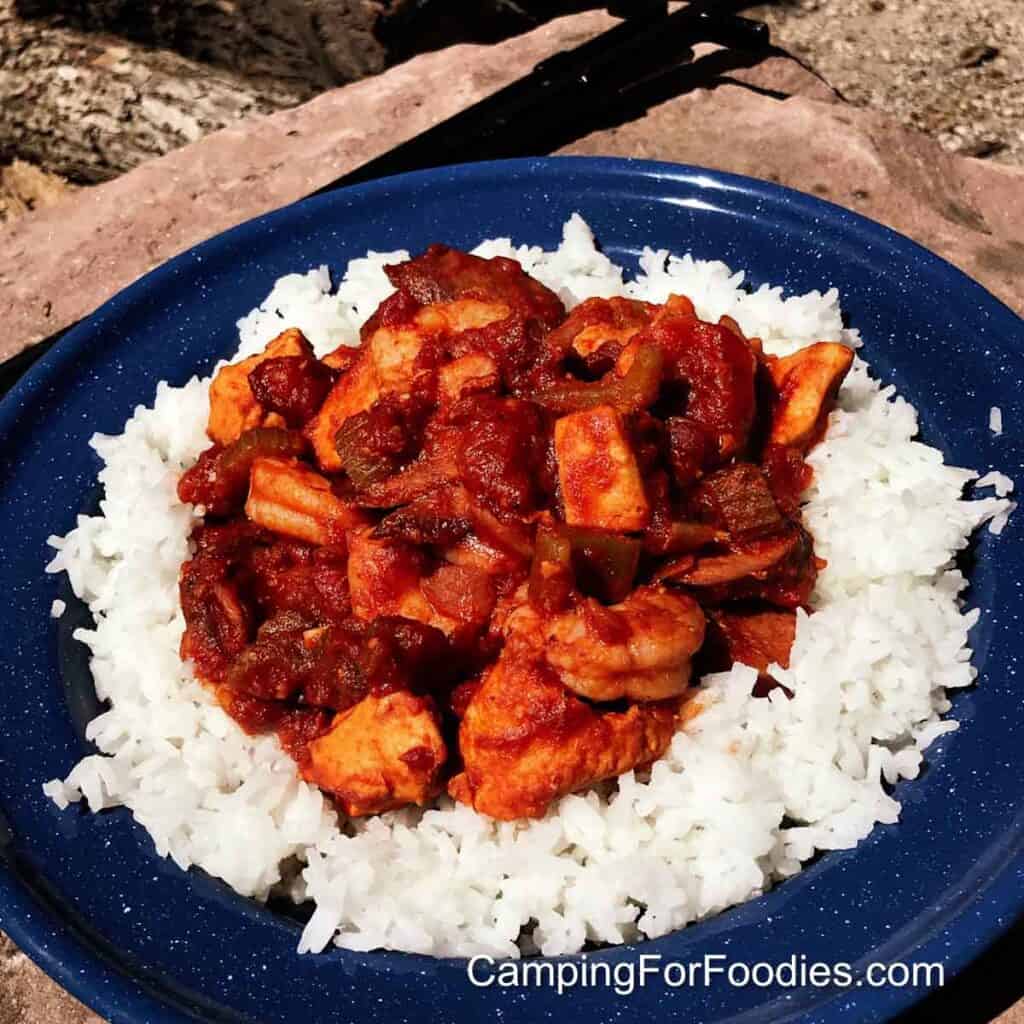 Easy Jambalaya Recipe by CampingForFoodies features a blue camping plate filled with rice and topped with a jambalaya Dutch oven dinner that includes shrimp, chicken and sausage in a deep red sauce.