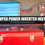 How To Install A Power Inverter In A Camper by CampingForFoodies features a blue DC-AC power adapter hung on a black bulkhead wall in an RV exterior storage compartment with wires, fuses, switches and a red tool box below. Text over the image reads camper power inverter install.