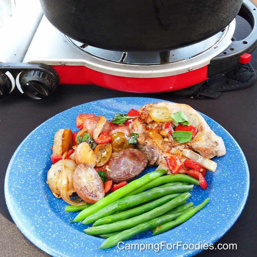 Pork Chops With Vinegar Peppers And Potatoes Recipe by CampingForFoodies features a blue camping plate with a golden-brown pork chop covered with sliced red cherry vinegar peppers, bell peppers, golden onion slices, multi-colored potatoes and bright green fresh basil with a Dutch oven sitting on a camp stove in the background.