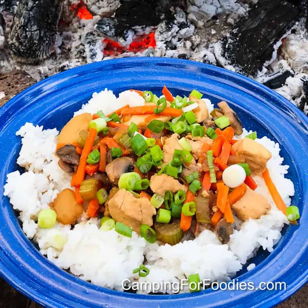 American Chinese Chop Suey by CampingForFoodies features a serving of chicken chop suey with carrots, mushrooms, celery and green onions over a bed of white rice on a blue camping plate with hot campfire coals in the background.