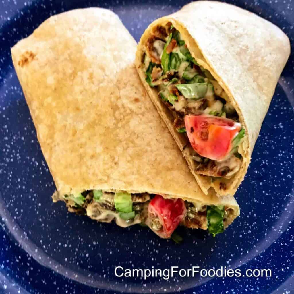 BLT Wraps For Camping by CampingForFoodies features a blue camping plate with a wheat tortilla wrap cut in half and stuffed with a mixture of green chopped salad, crispy bacon pieces and chopped tomatoes tossed in mayonnaise.