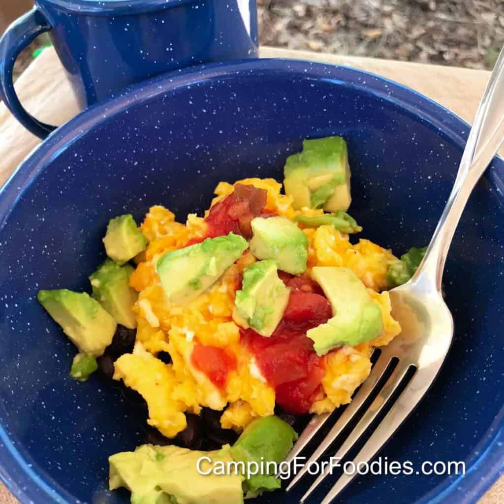 Breakfast Burrito Bowl by CampingForFoodies features a blue camping bowl filled with black beans, scrambled eggs, bright green avocado chunks and red salsa set next to a blue cup filled with camp coffee.