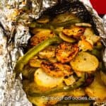 Camp Potatoes In Foil by CampingForFoodies features a foil packet filled with golden brown sliced potatoes, green bell pepper slices, seasoned with salt, pepper, garlic and butter.