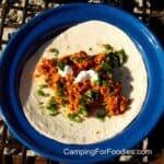 Camping Breakfast Burritos by CampingForFoodies features a sausage and egg breakfast burrito topped with sour cream and cilantro ready to be wrapped and folded on a blue camping plate set upon a campfire grill grate.