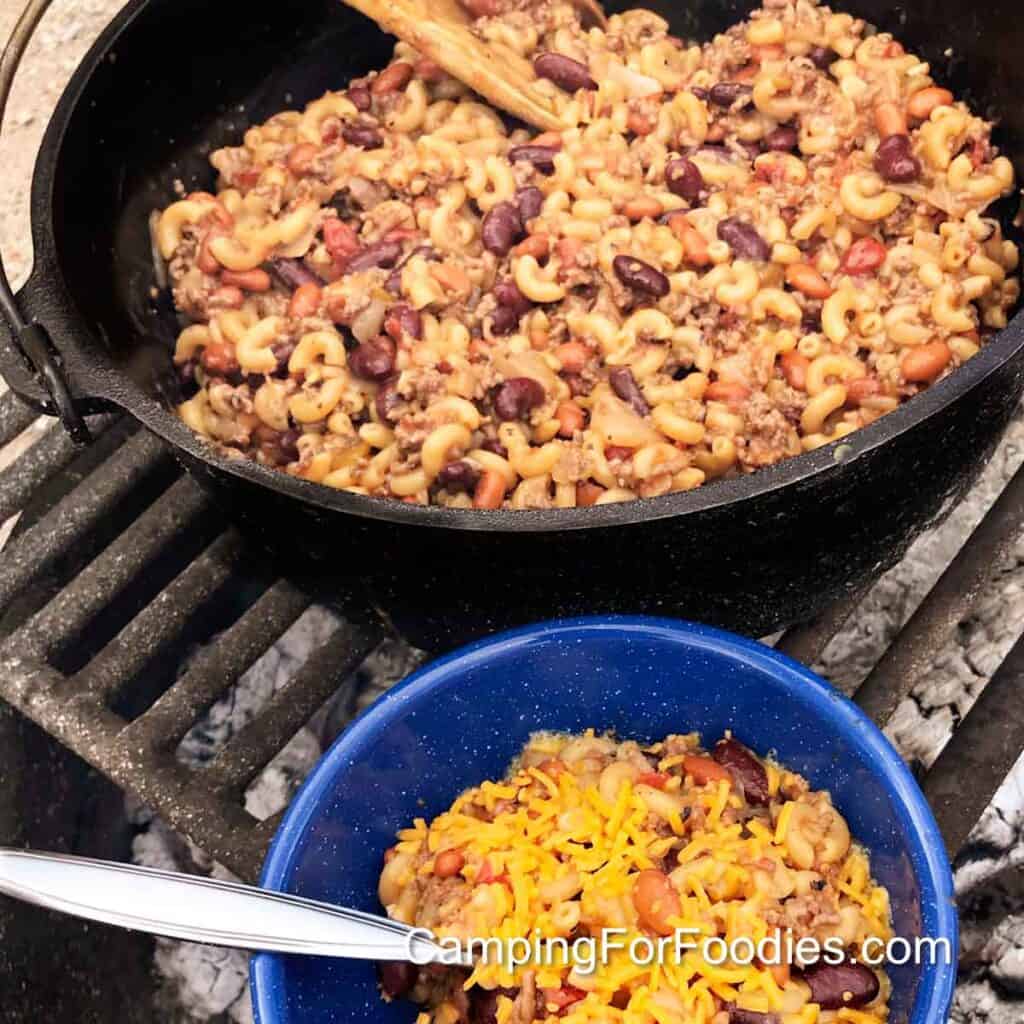 Camping Chili Mac by CampingForFoodies features a black cast iron pot set on a campfire cooking grate, filled with a one pot Dutch oven chili mac recipe with ground meat, red and brown beans, tomatoes and macaroni. A single serving is set aside in a blue camping bowl with yellow shredded cheddar cheese on top.