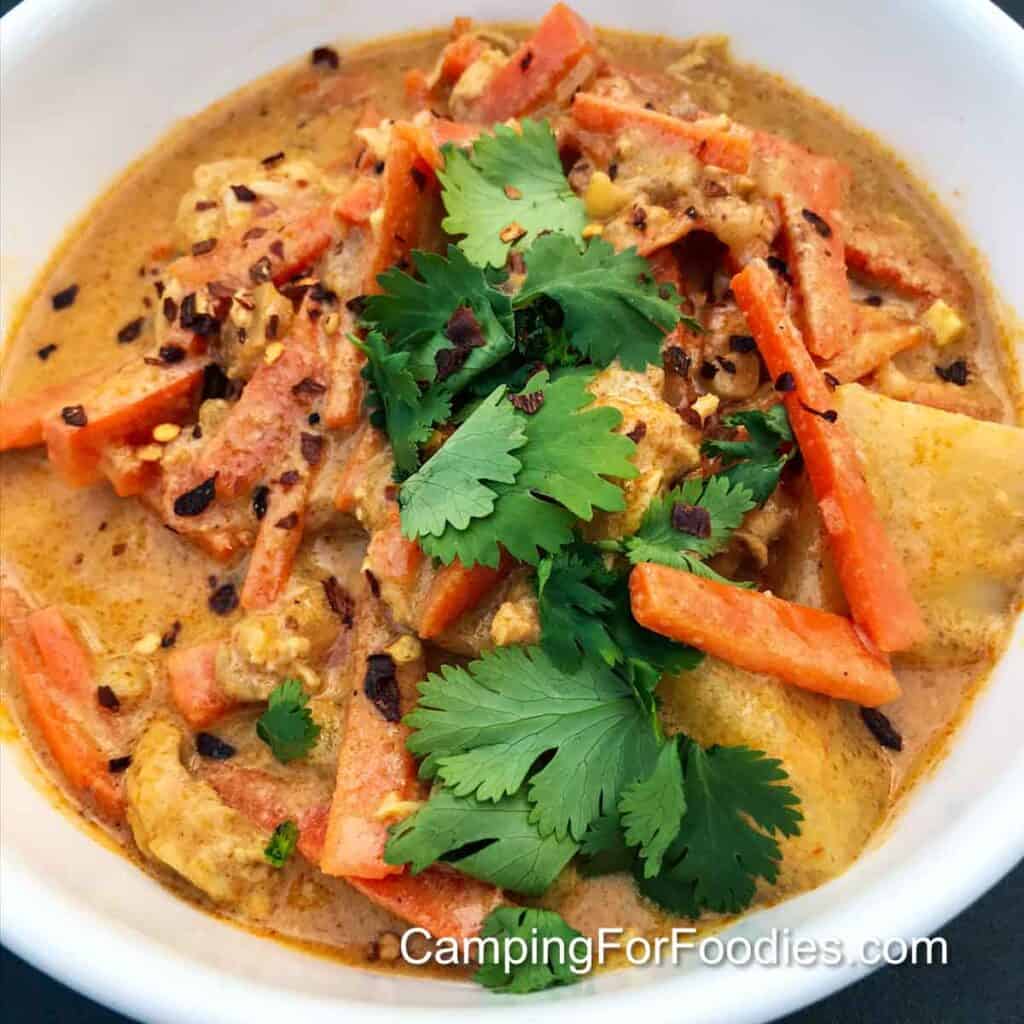 Camping Curry by CampingForFoodies features a white bowl filled with yellow and orange colored curry chicken topped with red and yellow hot pepper flakes and brightly colored greed cilantro.