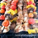 Camping Skewers by CampingForFoodies features skewers of charred grilled chicken cubes, cherry tomatoes, yellow bell peppers, onions and mushrooms sitting on a propane camping grill ready to be served.