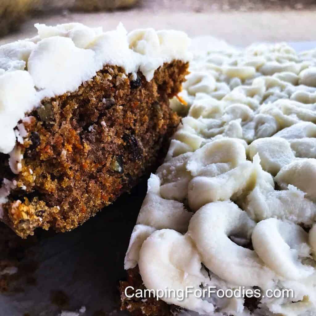 Carrot Cake In A Dutch Oven by CampingForFoodies features a moist carrot cake with raisins and nuts. Thick cream cheese frosting is piped in small swirls crowning the cake that is set on parchment paper. A slice is being lifted out and served.