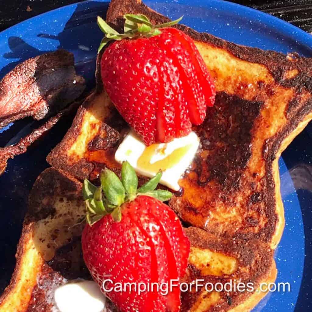 Cast Iron Campfire French Toast by CampingForFoodies features golden brown French toast topped with fresh strawberries, melting butter and real maple syrup on a blue camping plate.