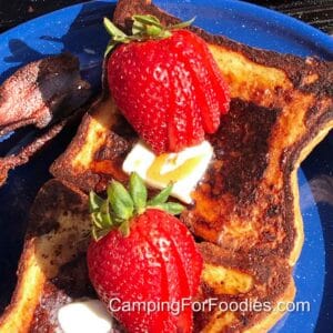 Cast Iron Campfire French Toast by CampingForFoodies features golden brown French toast topped with fresh strawberries, melting butter and real maple syrup on a blue camping plate.