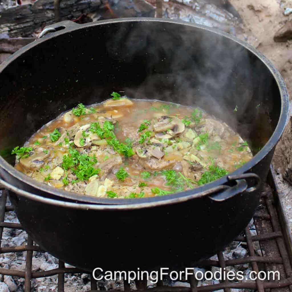 Dutch Oven Campfire Pork Chops by CampingForFoodies features a black cast iron Dutch oven set upon a campfire grill grate filled with golden brown pork chops and mushrooms in a brown gravy topped with bright green fresh parsley.