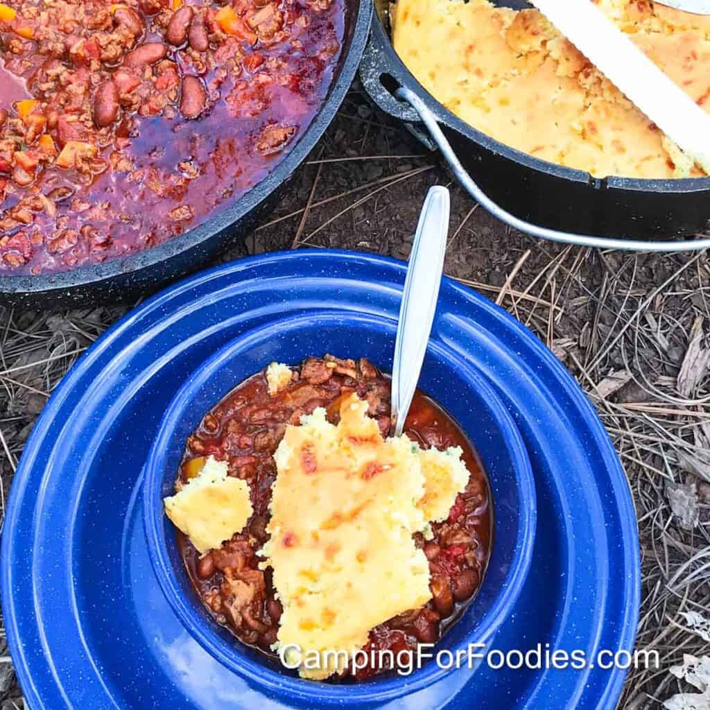 Dutch Oven Chili Recipe by CampingForFoodies features a blue camp bowl filled with beef chili topped with freshly made cornbread. In the background are two camp Dutch ovens, one filled with campfire chili, another filled with jalapeno cornbread.