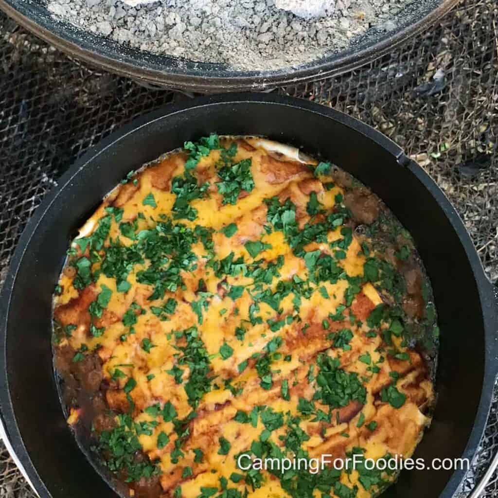Dutch Oven Enchiladas by CampingForFoodies features a black cast iron camp Dutch oven filled with chicken enchiladas baked in red sauce, topped with golden melted cheddar cheese and bright green chopped fresh cilantro.