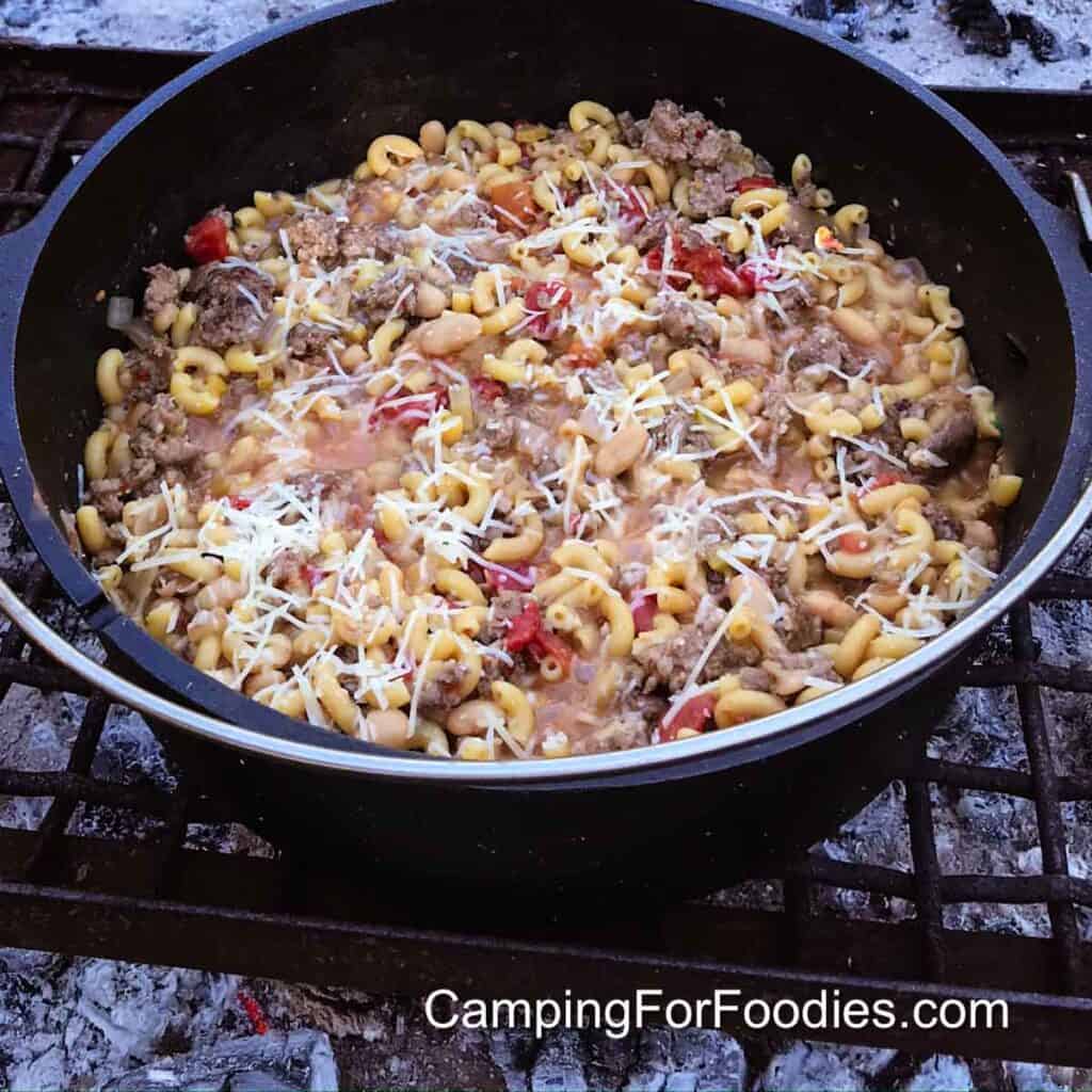 Dutch Oven Goulash by CampingForFoodies features a cast iron camp oven filled with goulash made with macaroni, meat, tomatoes and sprinkled with parmesan cheese. The pot is set on a grate in a campfire ring.