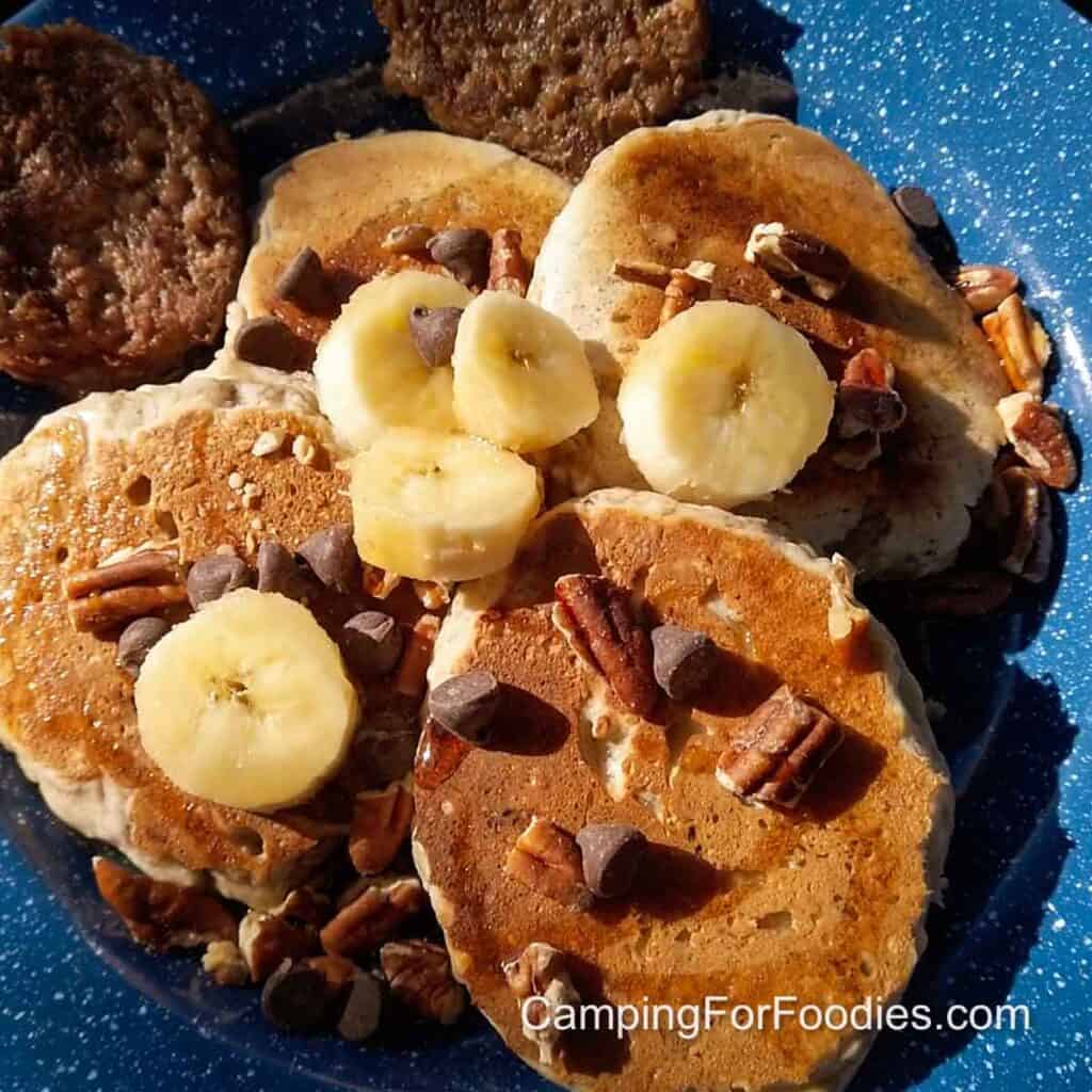 4 Ingredient Banana Pancakes by CampingForFoodies features golden brown pancakes topped with banana slices, nuts and chocolate chips on a blue camping plate.