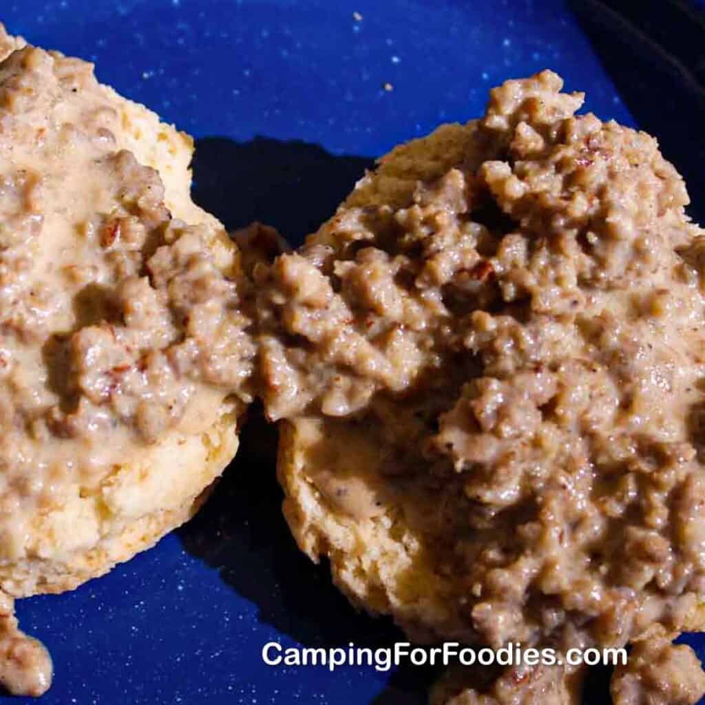 Camping Biscuits And Gravy by CampingForFoodies features homemade Dutch oven biscuits topped with sausage gravy in a thick sauce and set on a blue camping plate.