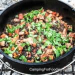 Dutch Oven Latin Chicken by CampingForFoodies features a black cast iron camp Dutch oven set in a campfire ring filled with cooked chicken, sweet potatoes, red bell pepper, black beans and bright green cilantro.