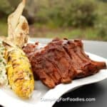 Dutch Oven Ribs Camping Recipe by CampingForFoodies features baby back pork ribs slathered in BBQ sauce on a white plate next to an ear of charred campfire corn and coleslaw side salad on a picnic table with a green forest in the background.