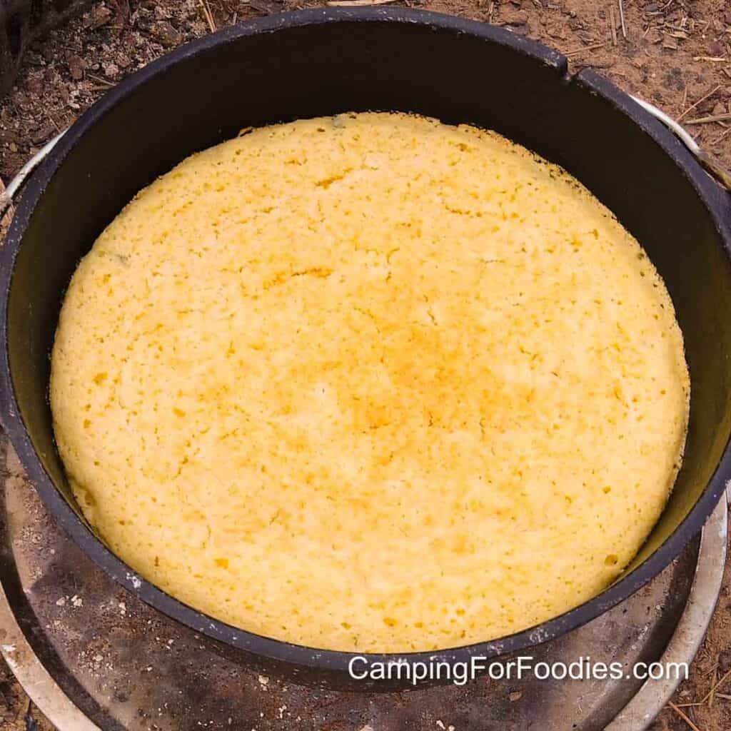 Campfire Cornbread by CampingForFoodies features a golden brown freshly-baked jalapeno and cornmeal bread in a black cast iron Dutch oven sitting on the ground at a campsite with brown pine needles in the background.