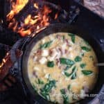 Hot Italian Sausage Campfire Soup by CampingForFoodies features a black cast iron camp Dutch oven filled with creamy sausage and potato soup with browned bacon and fresh green spinach. The camping soup pot is set on a campfire grate with orange and red flames in the background at the bottom of a fire ring.