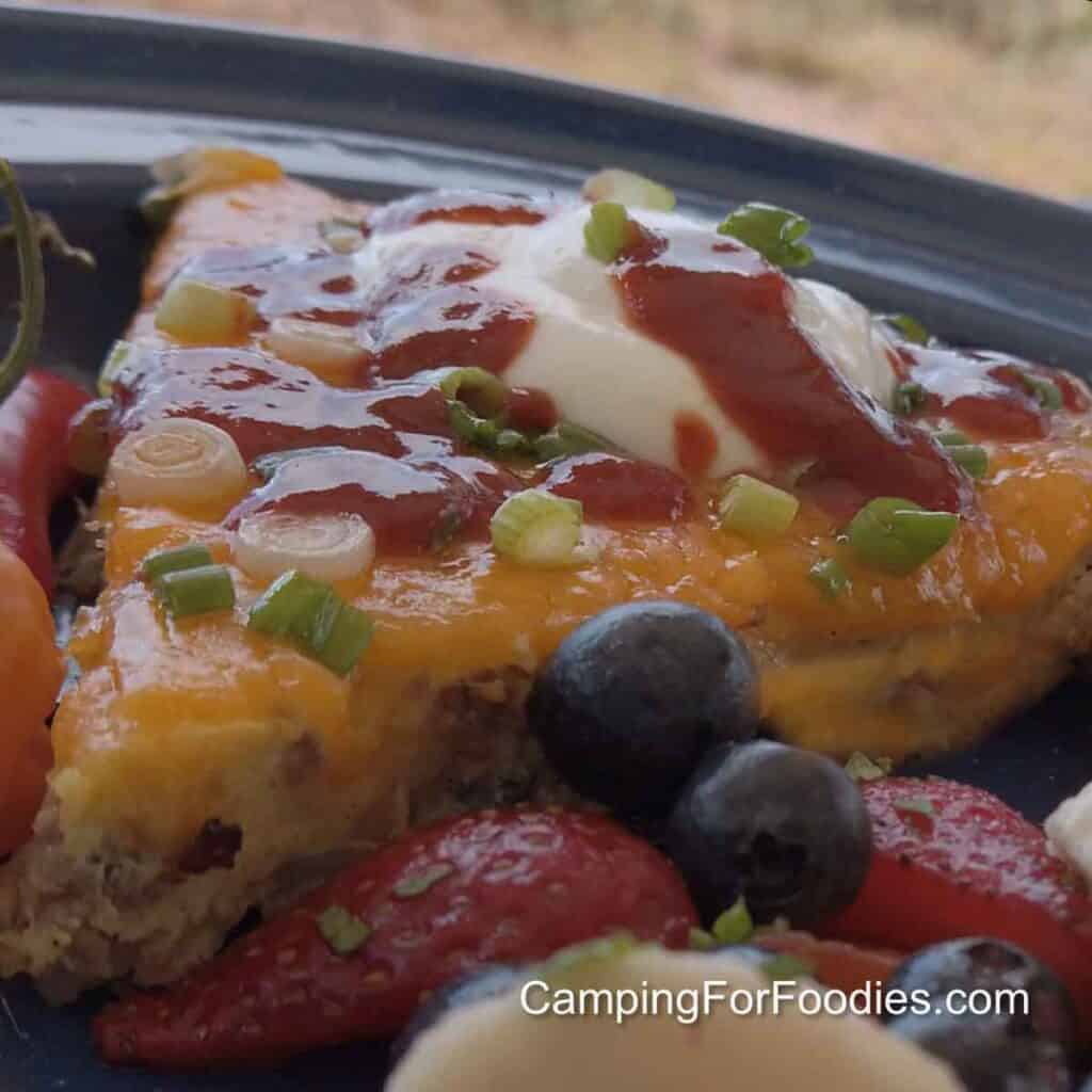 Mountain Man Breakfast Recipe by CampingForFoodies features a slice of a sausage, egg, hashbrown and golden melted cheese casserole on a blue camping plate. The meal is topped with sliced green onions, sour cream and red hot sauce. Fresh blueberries, sliced strawberries and bananas are served on the side. Orange and red fresh peppers are also on the plate.