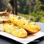 Campfire Corn On The Cob by CampingForFoodies features three ears of charred sweet corn on the cob with slightly charred husks pulled away from the cobs and stacked on a white plate. Basil butter is melting over the hot corn set outside on a sunny day with a dense green forest in the background.