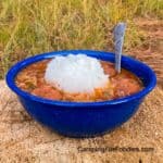 Campfire Dutch Oven Gumbo by CampingForFoodies features a blue camping bowl filled with chicken, sausage and shrimp gumbo in a brownish red sauce and a scoop of white rice with a spoon in the bowl that is set on a chopped firewood log with green meadow grass in the background.