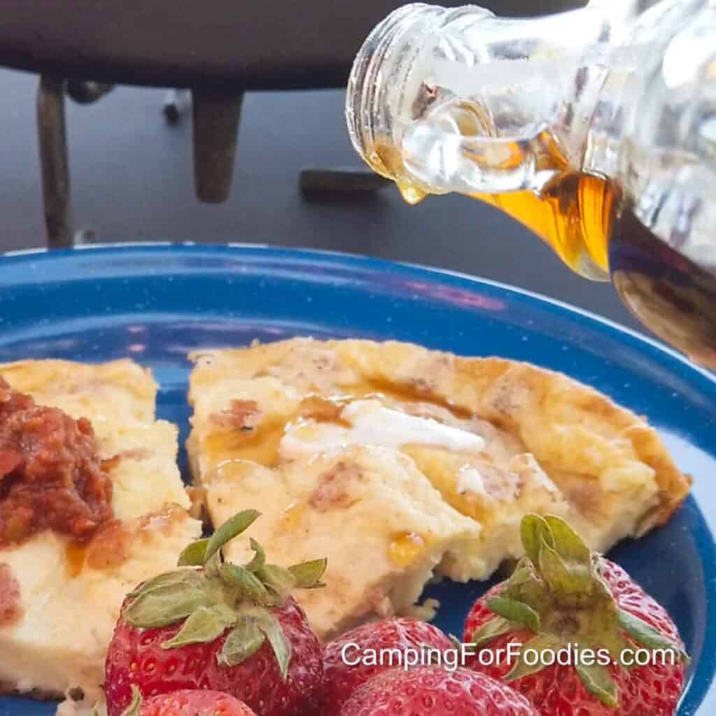 Swedish Oven Pancake by CampingForFoodies features two slices of a golden-brown, oven-baked puffed pancake on a blue camping plate. One slice is topped with red salsa for a savory version of the recipe, the other slice is topped with melting butter and maple syrup. A glass syrup bottle is over the plate glistening in sunshine. Bright red strawberries garnish the bottom of the plate.