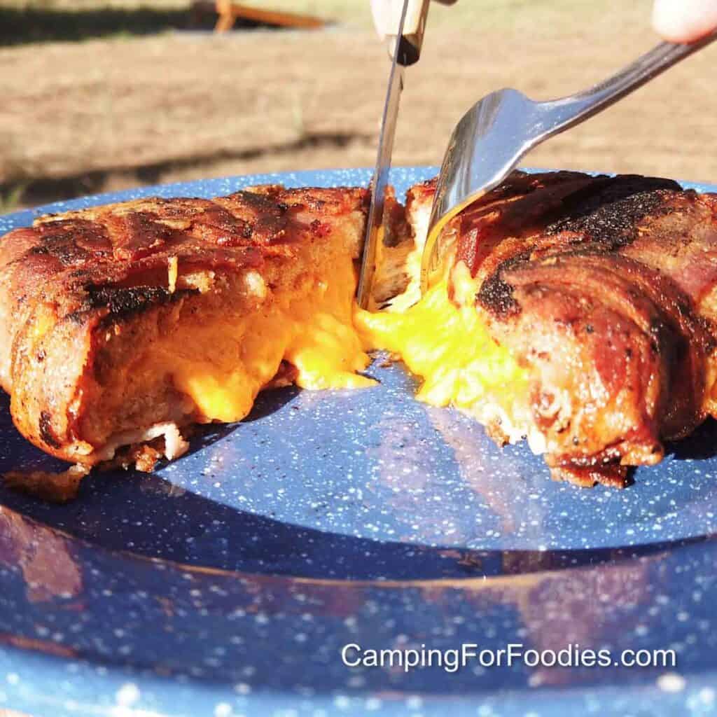 Bacon Wrapped Grilled Cheese by CampingForFoodies features golden brown crispy bacon wrapped around a grilled cheese sandwich that is set on a blue camping plate. A fork and knife are being used to slice the sandwich as golden yellow cheddar cheese oozes from the center of the sandwich onto the plate.