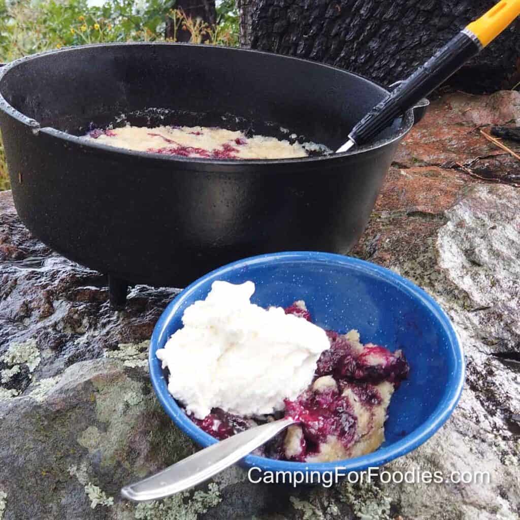 Campfire Dutch Oven Blueberry Cobbler by CampingForFoodies features a blue camping bowl with a serving of blueberry cobbler and vanilla ice cream set on a large rock under a tree. A black cast iron camp Dutch oven containing the rest of the baked cobbler is set behind the bowl that is ready to be served.