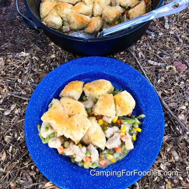 Dutch Oven Chicken Pot Pie by CampingForFoodies features a blue camping plate filled with a big helping of chicken pot pie dinner in front of a cast iron camp Dutch oven with a serving spoon and a fluffy biscuit upper-crust chicken pot pie with carrots, peas, green beans and corn.