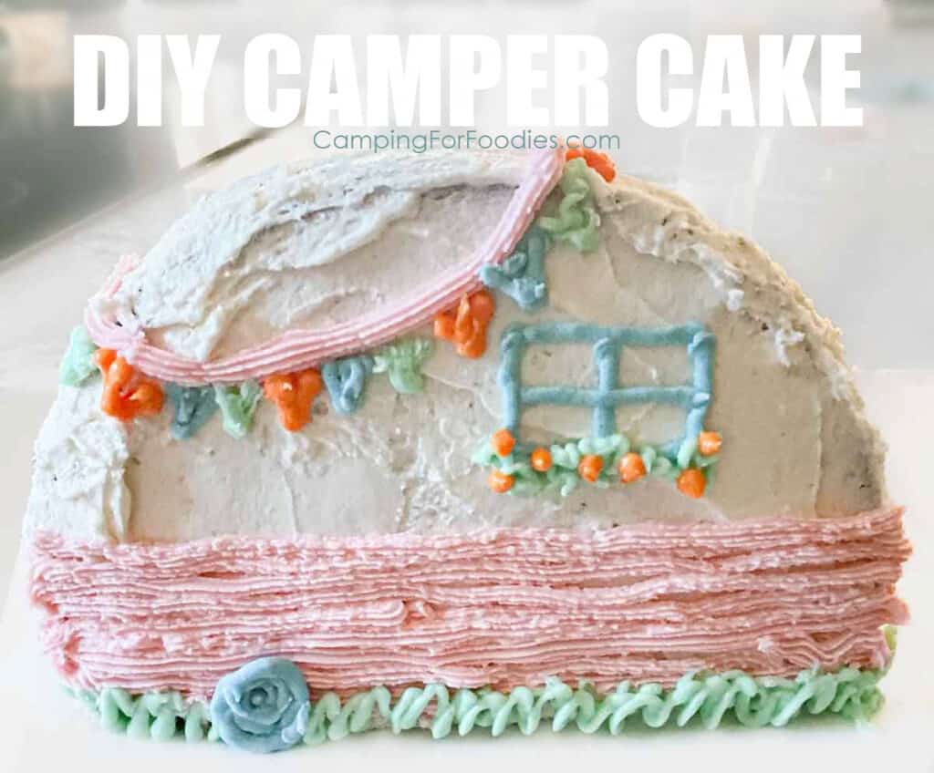 DIY Camp Theme Cake by CampingForFoodies features a homemade cake decorated with white buttercream frosting. The cake is standing upright and made to look like a retro RV trailer with a blue tire and window with window box filled with orange flowers, a pink strip around the body of the camper and blue, orange and green flags hanging around the outside of the roof. The trailer is sitting on green icing to look like grass. Text over the image reads DIY Camper Cake.