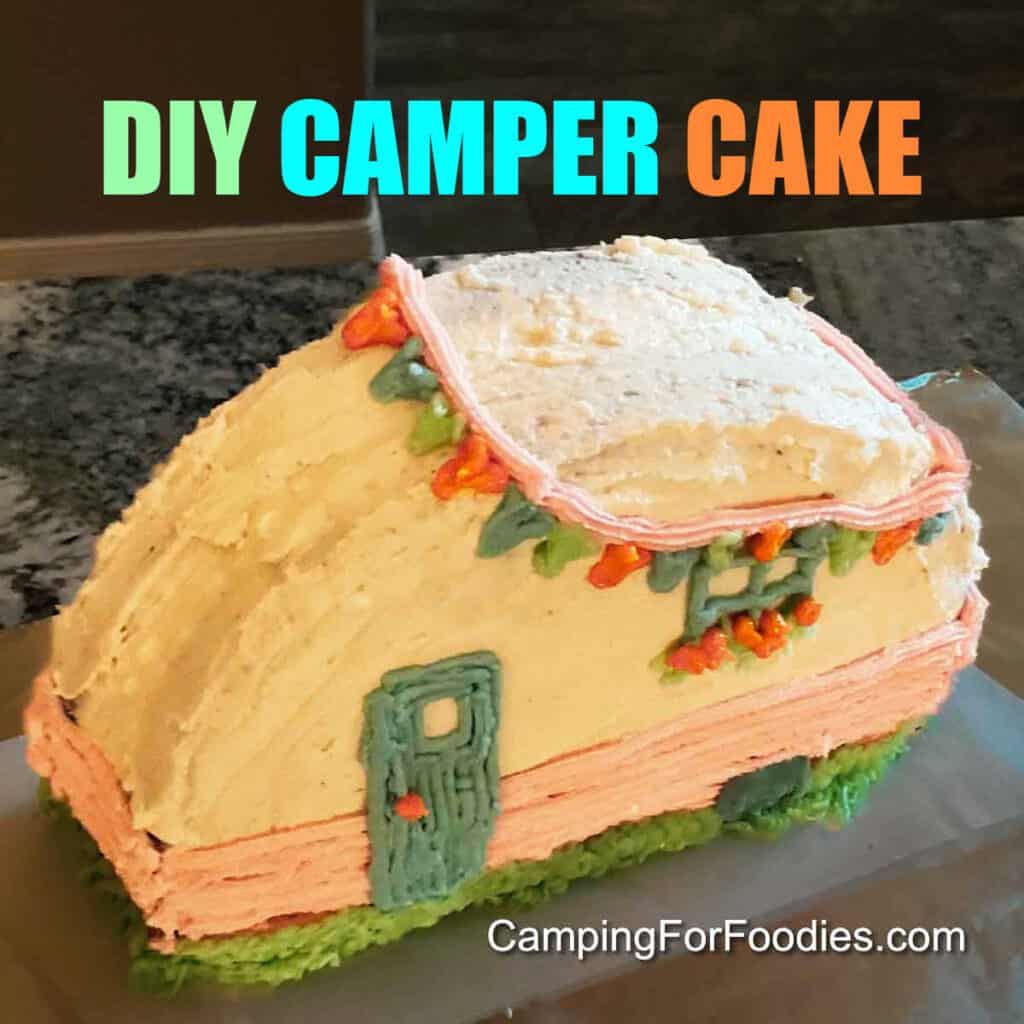 DIY Camper Cake by CampingForFoodies features a homemade cake decorated with white buttercream frosting. The cake is standing upright and made to look like a retro RV trailer with a blue door, window with window box filled with orange flowers, a pink strip around the body of the camper and blue, orange and green flags hanging around the outside of the roof.