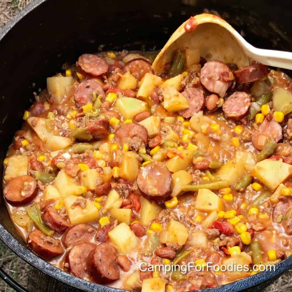 Dutch Oven Cowboy Stew by CampingForFoodies features a black cast iron camp Dutch oven sitting on the ground filled with a sauce and browned ground beef, andouille sausage, potatoes and canned corn, tomatoes, green beans and green chilies.