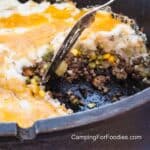 Dutch Oven Shepherd's Pie by CampingForFoodies features a black cast iron Dutch oven filled with a layered casserole containing browned ground beef, mixed vegetables, a layer of mashed potatoes and melted cheddar cheese. A spatula sits in the pot ready to scoop out the next serving.