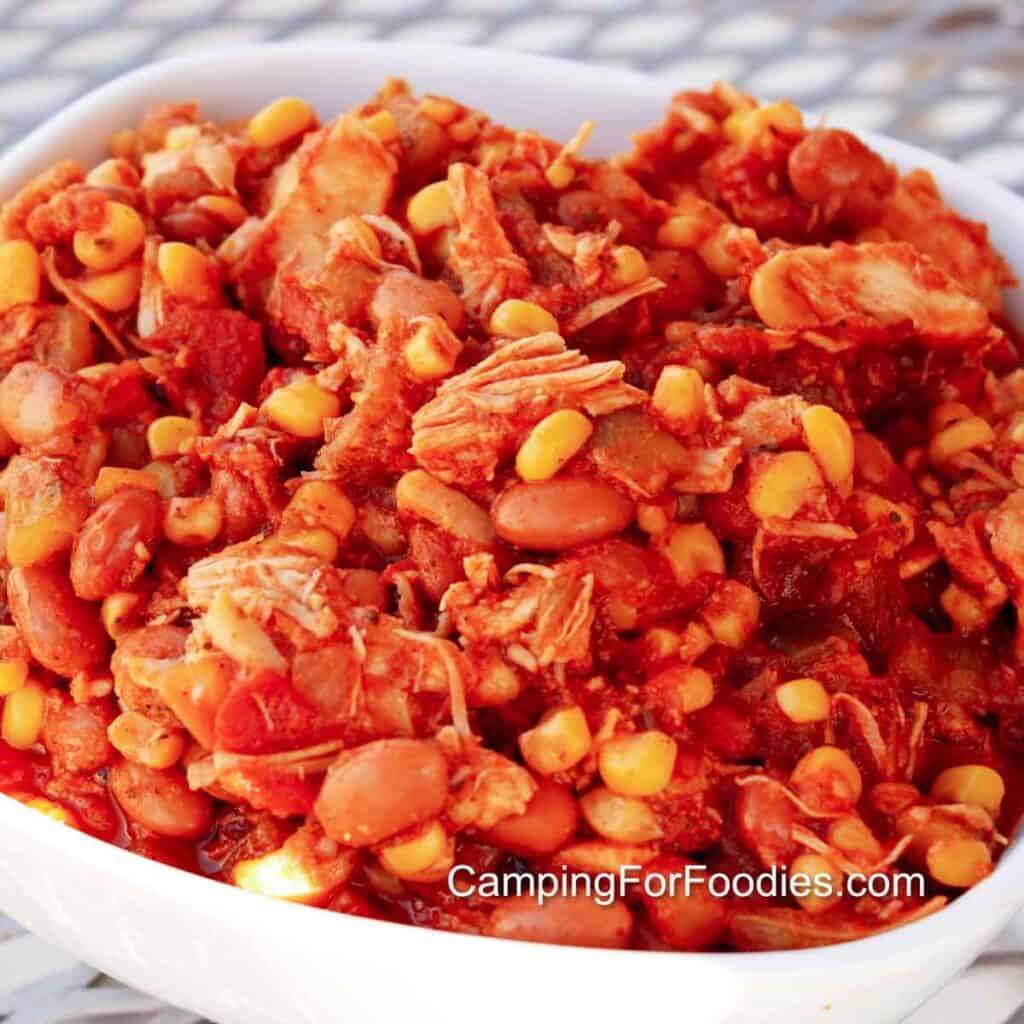 5 Can Camp Chili by CampingForFoodies features a white serving bowl filled with canned chicken meat that is covered in a red sauce from heating the meat with canned Mexican style corn, pinto beans, green chilies and salsa.