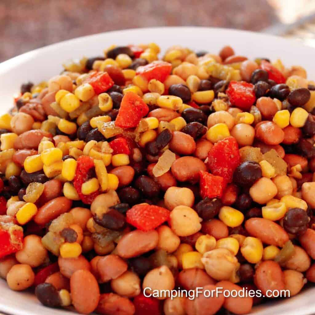 Mexican Three Bean Salad by CampingForFoodies features a white serving bowl filled with canned garbanzo, black and pinto beans, yellow kernels of canned Mexican style corn, green chilies and red peppers,