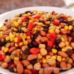 Mexican Three Bean Salad by CampingForFoodies features a white serving bowl filled with canned garbanzo, black and pinto beans, yellow kernels of canned Mexican style corn, green chilies and red peppers,