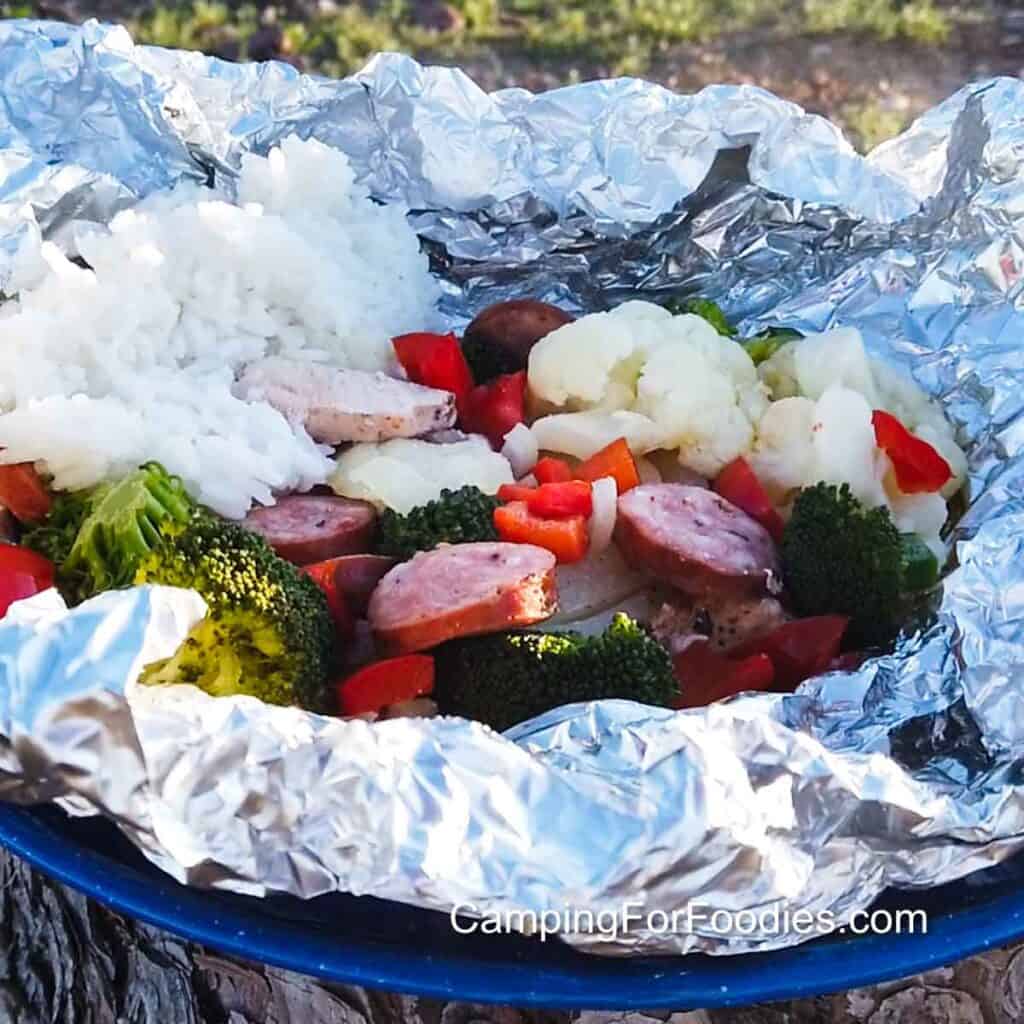 Cajun Chicken Foil Packets by CampingForFoodies features a silver-colored piece of tinfoil used as a bowl to hold a serving of Cajun chicken. The pieces of white cauliflower, green broccoli, red bell pepper and white onion slices compliment the chicken and andouille sausage pieces. A serving of white rice is behind the food and the foil packet is set on a blue camping plate with green shrubbery in the background.